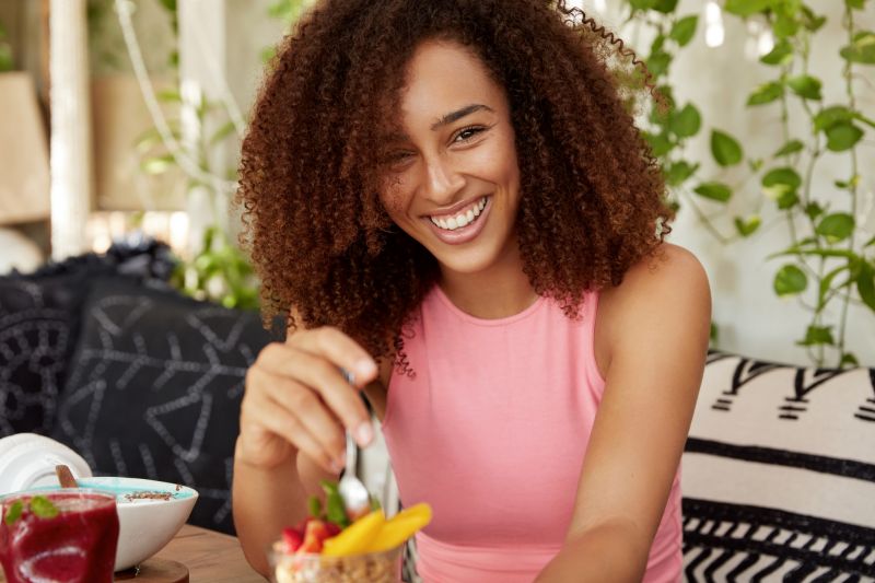 mulher-encaracolada-de-expressao-alegre-come-deliciosa-sobremesa-esta-de-bom-humor-passa-o-tempo-em-aconchegante-cafeteria-aprecia-saborosa-salada-de-frutas-mulher-atraente-descansando-sozinha-apos-a-excursao